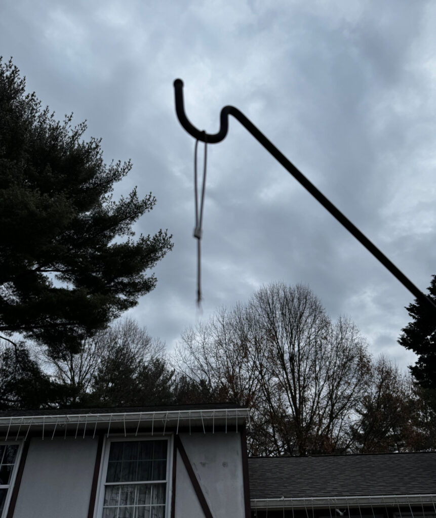 A photo looking up at the hook for a birdfeeder. A broken hanger wire is dangling from the hook.  In the background we see leafless trees and cloudy skies.