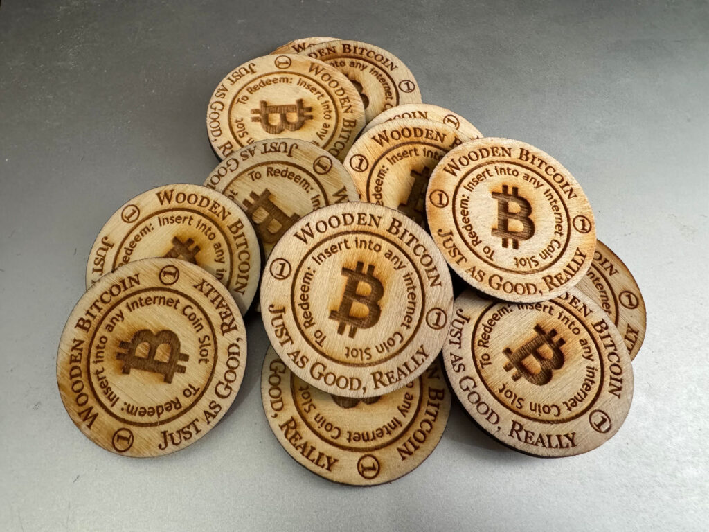 A small pile of wooden disks on a silver background. An outer circle on each disk reads "Wooden Bitcoin. Just as Good, Really." Text on an inner circle reads "To Redeem: Insert into any Internet Coin Slot." The Bitcoin logo appears at the center of each disk.