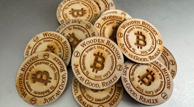 A small pile of wooden disks on a silver background. An outer circle on each disk reads "Wooden Bitcoin. Just as Good, Really." Text on an inner circle reads "To Redeem: Insert into any Internet Coin Slot." The Bitcoin logo appears at the center of each disk.