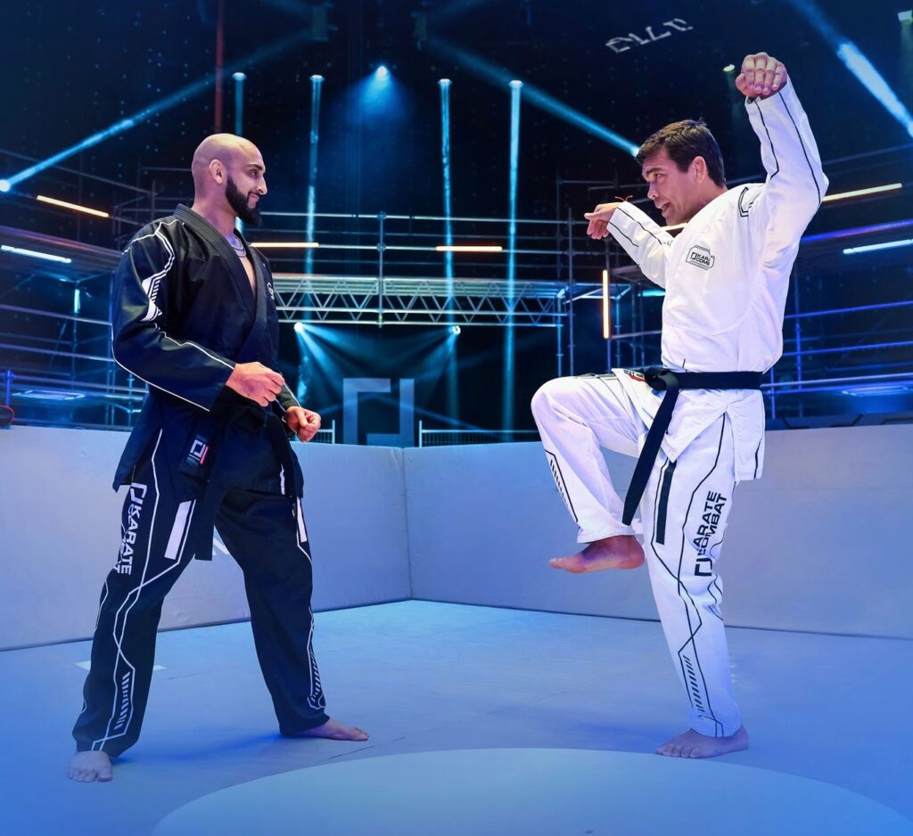 Two men in martial arts uniforms, standing on a mat in what appears to be an arena or gymnasium.

*   **Two Men**
    *   The man on the left is bald and has a beard.
        *   He is wearing a black martial arts uniform with white accents.
        *   His hands are clasped together in front of him.
    *   The man on the right has short brown hair.
        *   He is wearing a white martial arts uniform with black accents.
        *   His left leg is raised, and his arms are bent at the elbows.

The image suggests that the two men are engaged in some kind of martial arts training or competition.